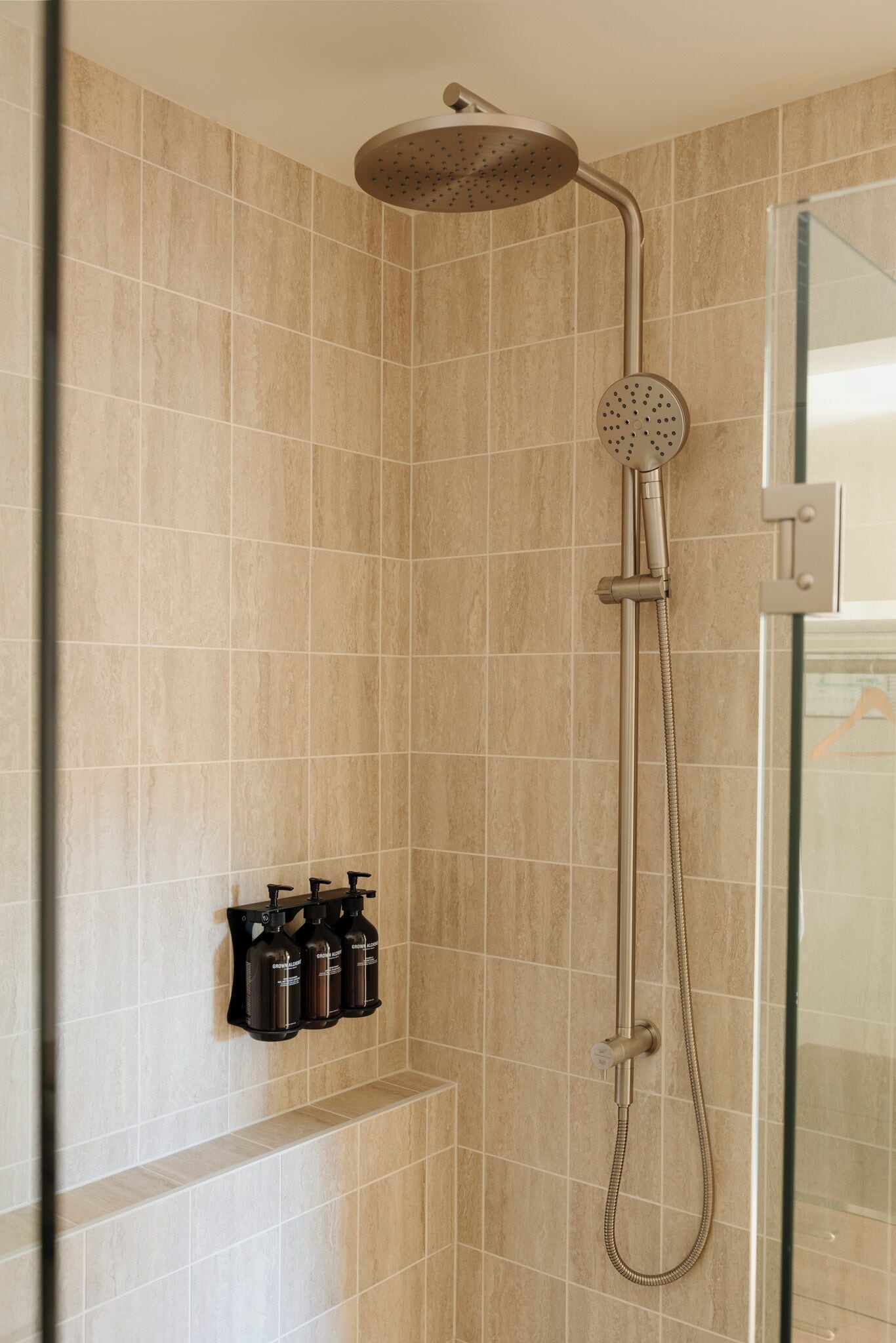 Ducha con cabezal de ducha manual y grifo empotrado en la pared. Hay tres botellas de champú o acondicionador en un estante debajo de la ducha.