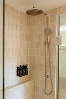 Ducha con cabezal de ducha manual y grifo empotrado en la pared. Hay tres botellas de champú o acondicionador en un estante debajo de la ducha.