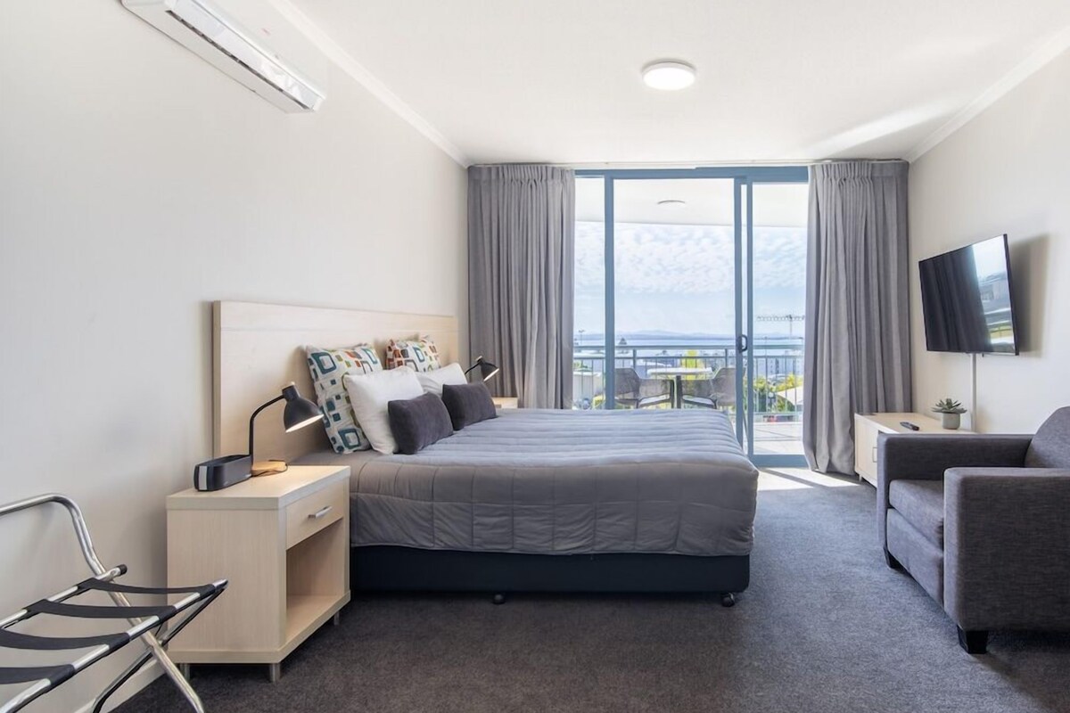 A hotel room with a bed, a desk, a chair, a TV, and a balcony with curtains.