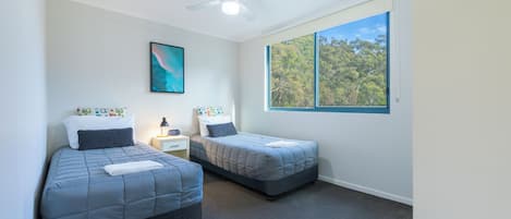 Quarto com duas camas, um ventilador de teto, uma mesinha de cabeceira, uma luminária e uma janela com vista para árvores.