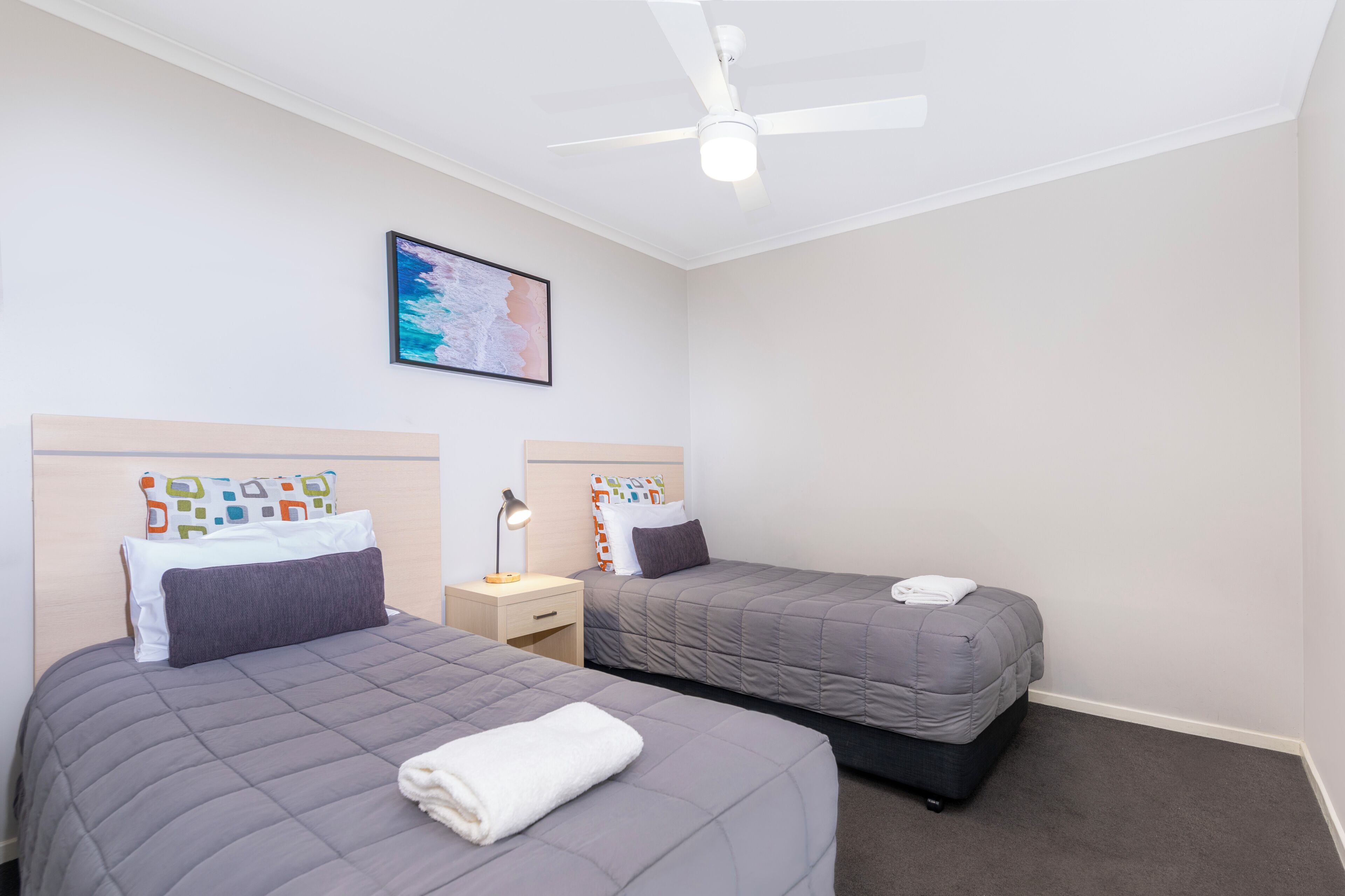 Une chambre d’hôtel avec deux lits, un ventilateur de plafond, une table de chevet avec une lampe et un tableau encadré au mur.