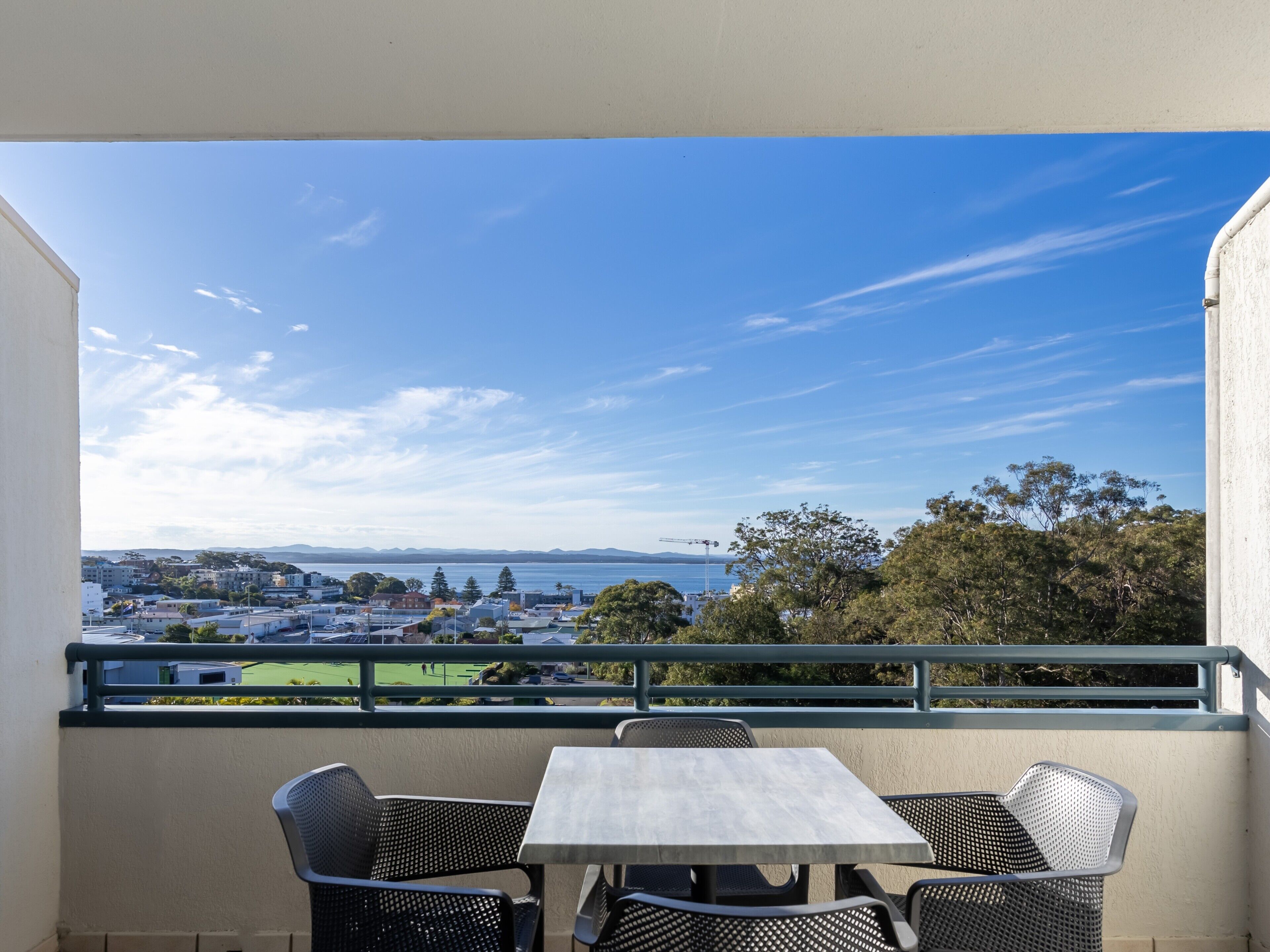 A balcony with a table and chairs, overlooking a coastal town with buildings and trees.