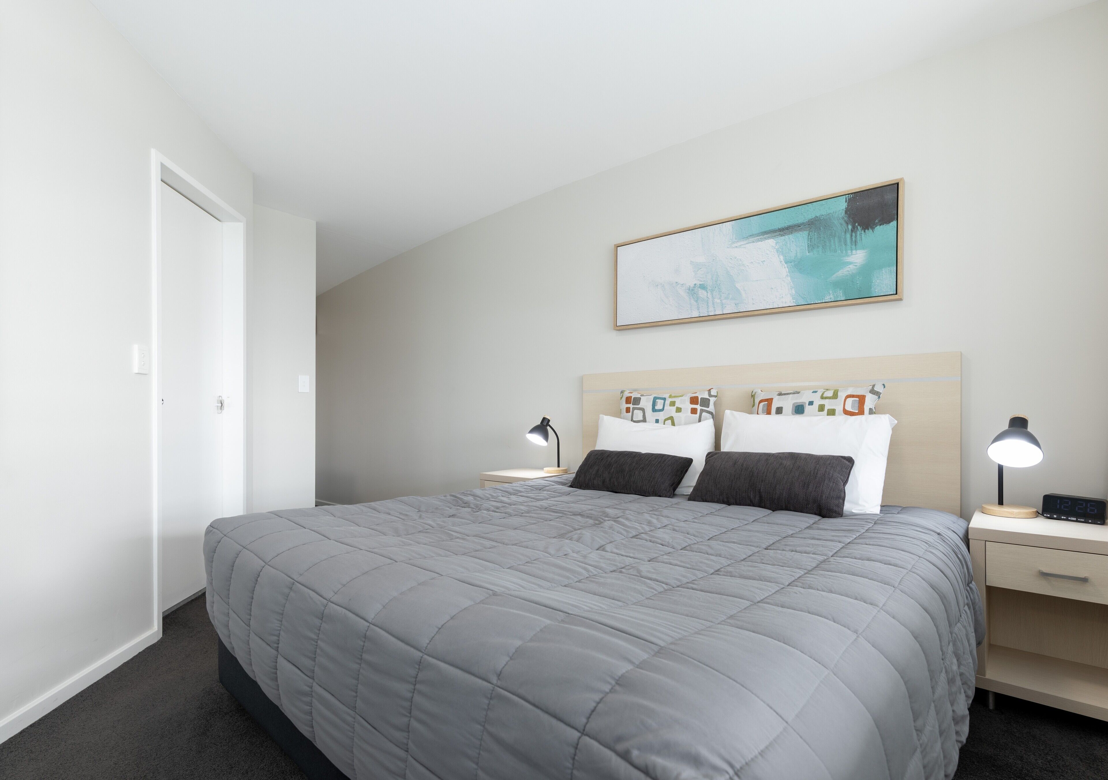 A neatly arranged hotel room with a large bed, two bedside tables with lamps, a wall-mounted painting, and a door to the left.