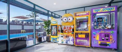 Un espacio interior moderno con paredes de vidrio, una piscina y coloridas máquinas de arcade, incluyendo juegos con temática de 'Toy Story' y 'Chocolate Factory'.