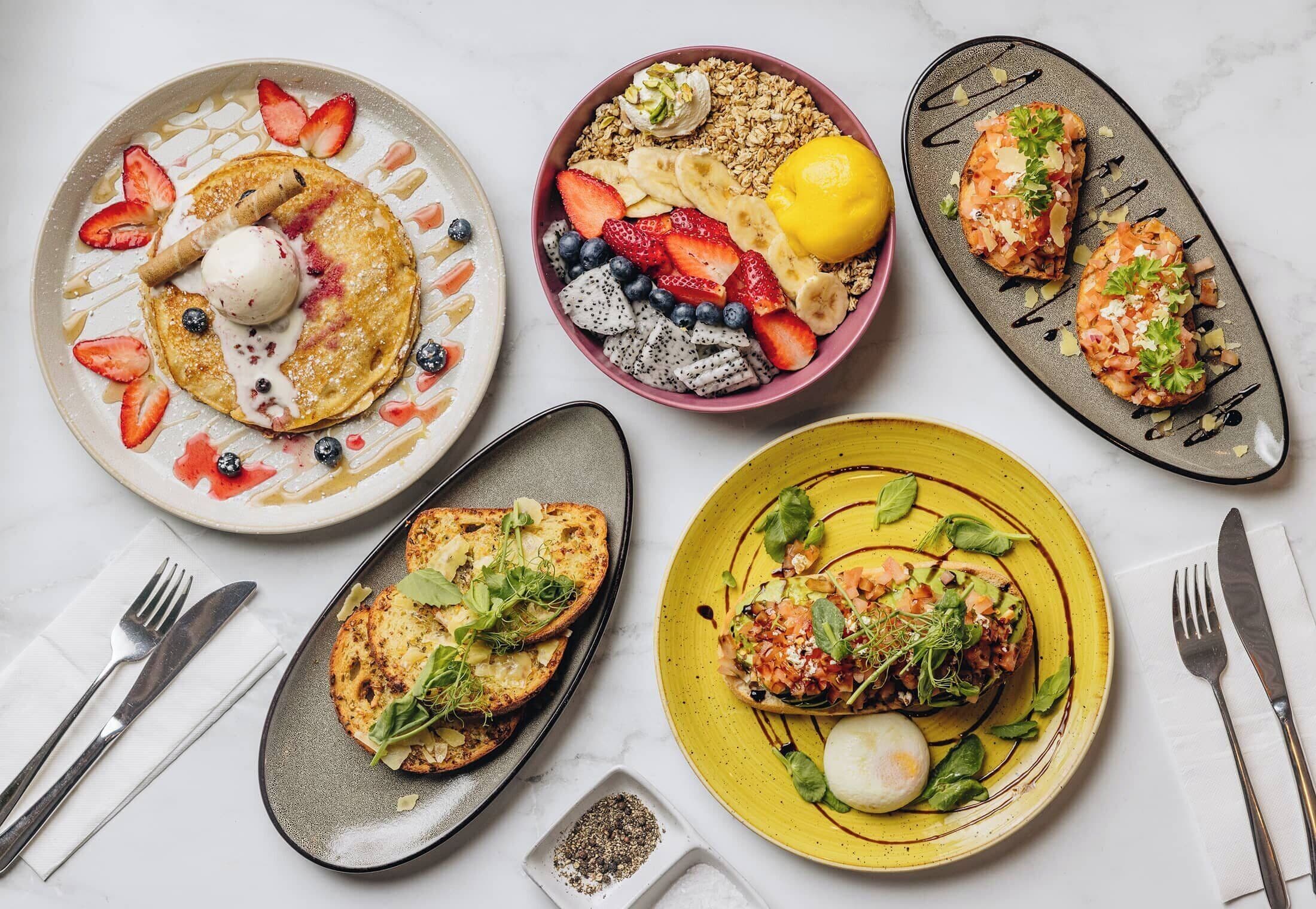 Diverse pietanze per la colazione su piatti, tra cui pancake, una ciotola di frutta e granola, e un piatto con uovo in camicia e verdure.