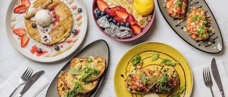 Diverse pietanze per la colazione su piatti, tra cui pancake, una ciotola di frutta e granola, e un piatto con uovo in camicia e verdure.