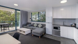 Uma sala de estar moderna com sofá, puf e mesa de centro, área de jantar com cadeiras, cozinha equipada e varanda com vista para árvores.