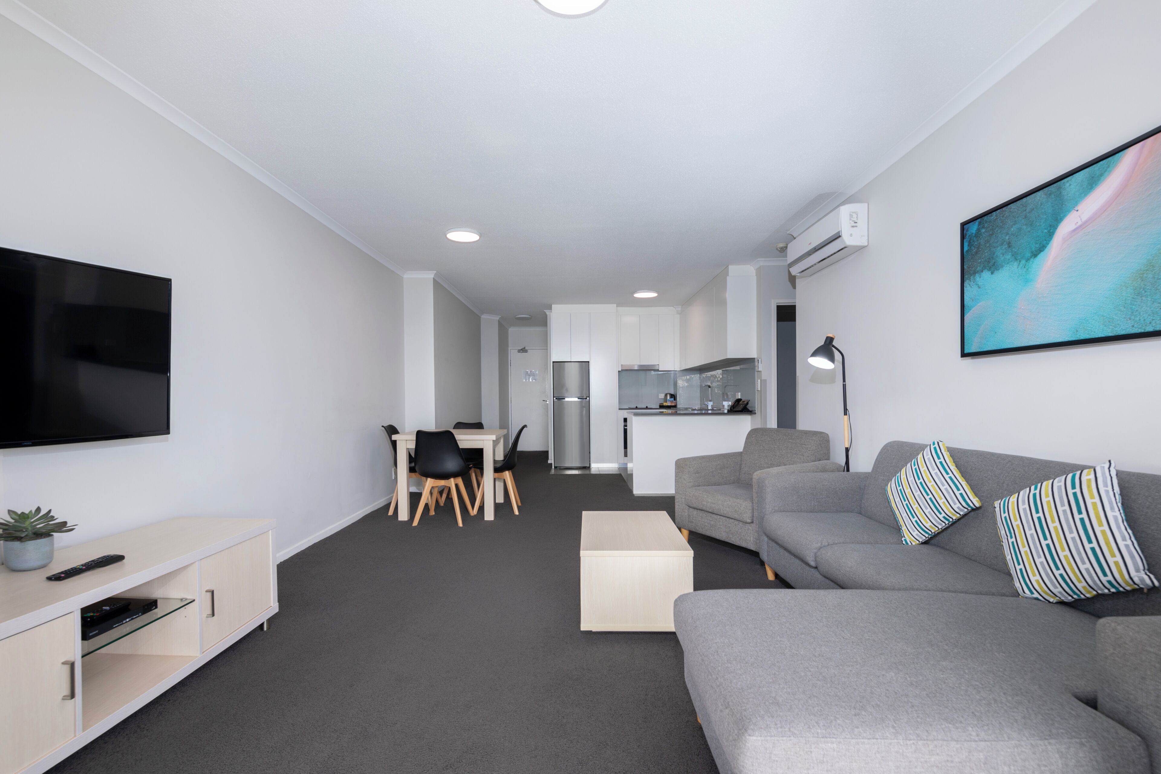 A modern living room with a grey sofa, a flat-screen TV, a dining area with a table and chairs, and a kitchen in the background.