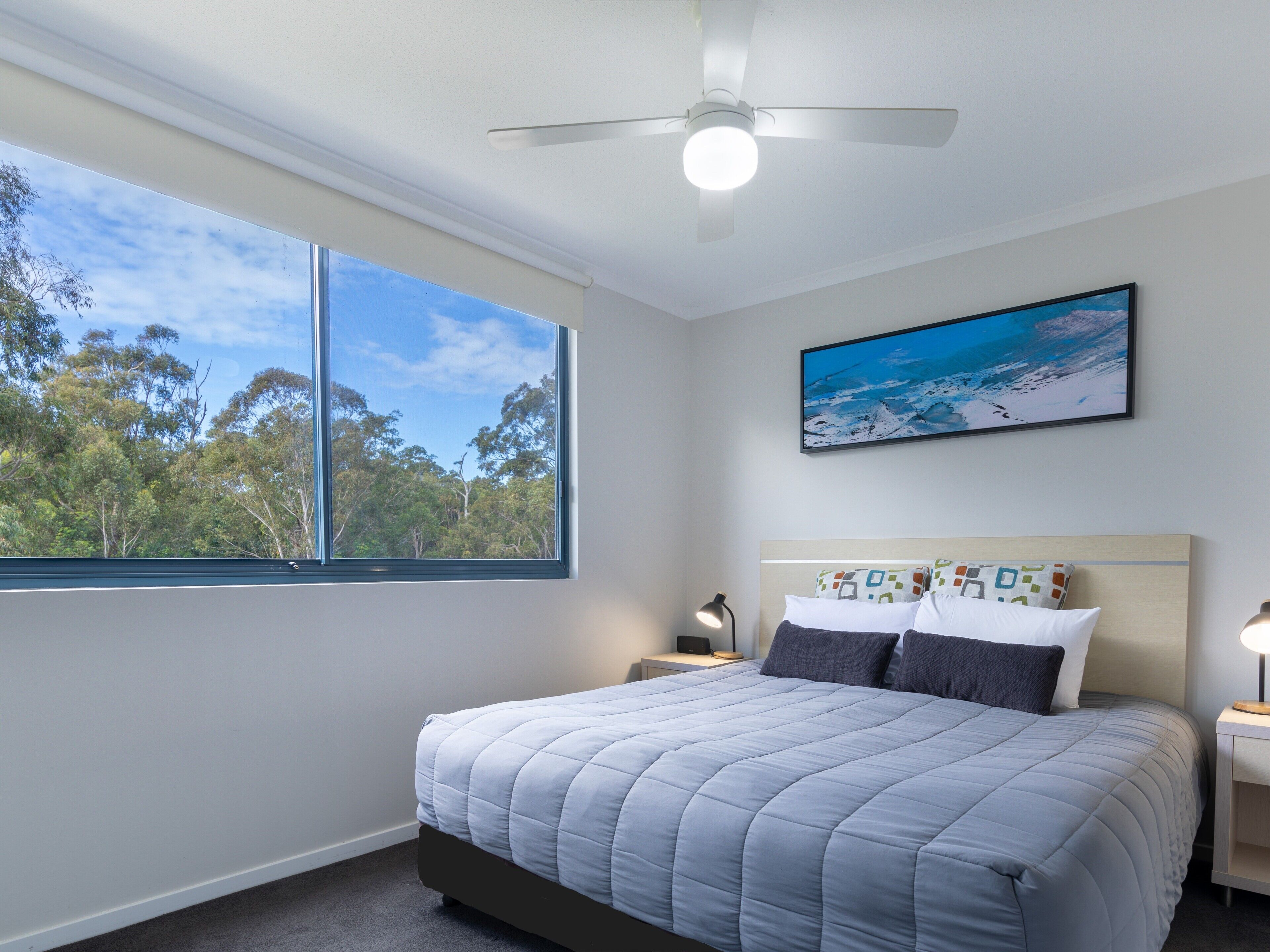 A bedroom with a bed, a ceiling fan, a window with a view of trees, and a painting on the wall.