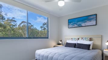 Un dormitorio con cama, ventilador de techo, ventana con vista a árboles y un cuadro en la pared.