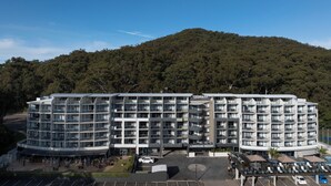 Hotel de vários andares com varandas, estacionamento e vista para uma colina arborizada.