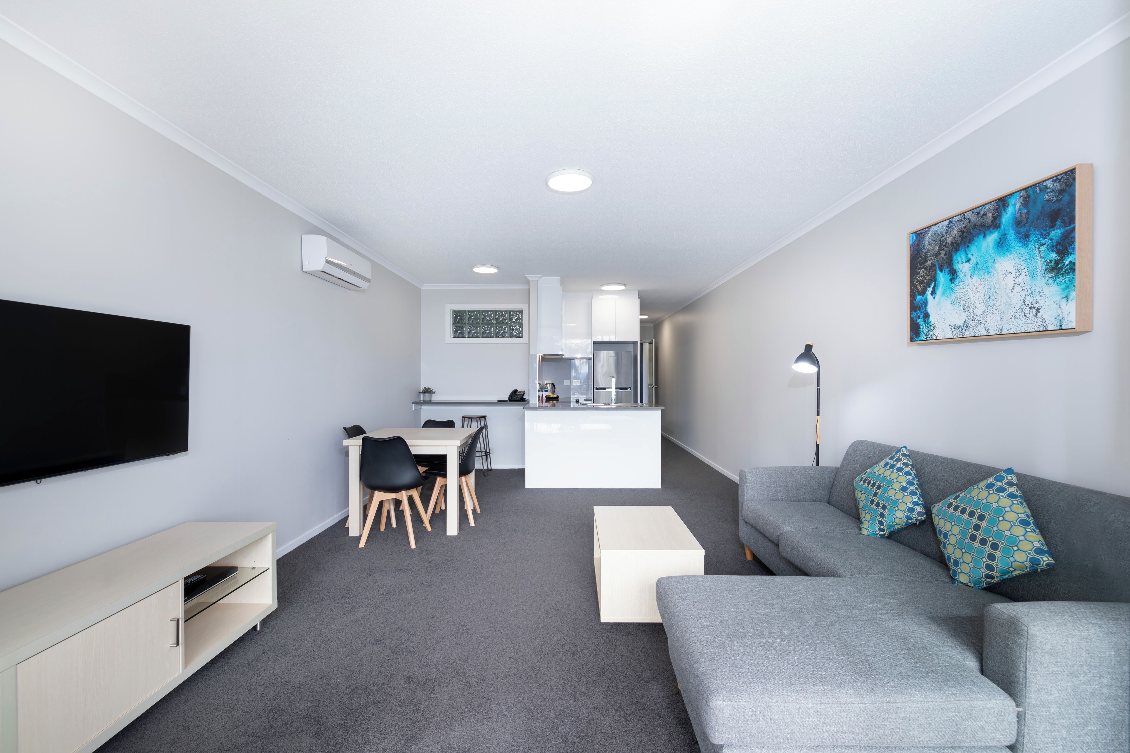 Un salon moderne avec un canapé gris, un téléviseur à écran plat, un coin repas avec des chaises et une cuisine en arrière-plan.