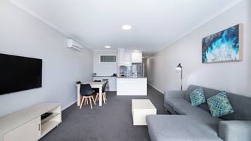 Una sala de estar moderna con un sofá gris, un televisor de pantalla plana, un comedor con sillas y una cocina al fondo.
