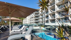Uma área à beira da piscina com espreguiçadeiras e guarda-sóis, uma piscina e um edifício com várias andares e varandas.