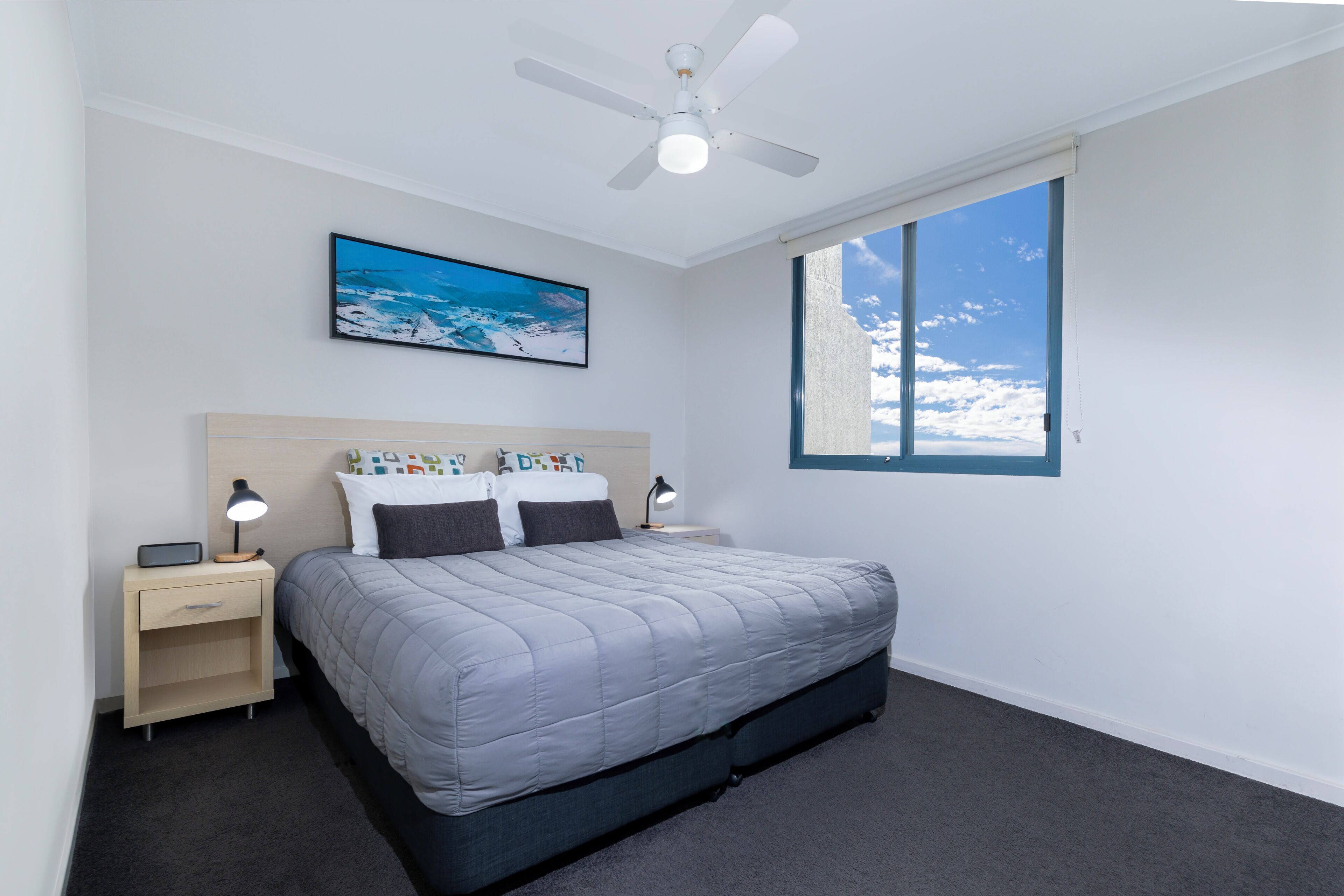 A bedroom with a bed, nightstands, a ceiling fan, a window with a view of the sky, and a painting on the wall.