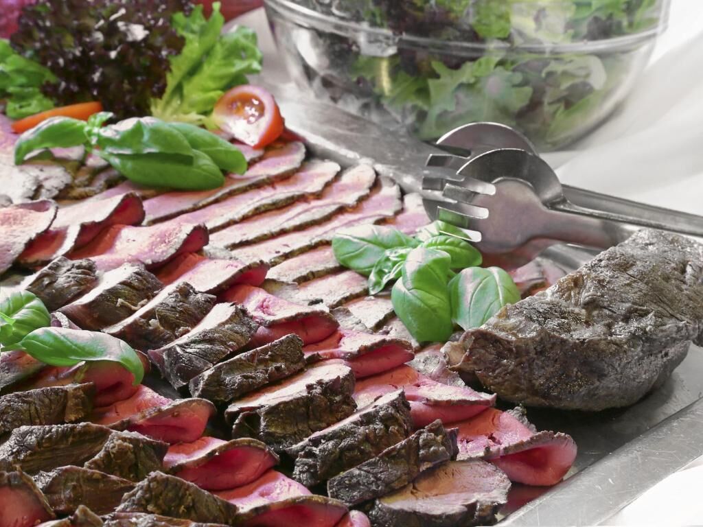 A platter of sliced steak garnished with basil leaves, accompanied by a fork and a large rock.
