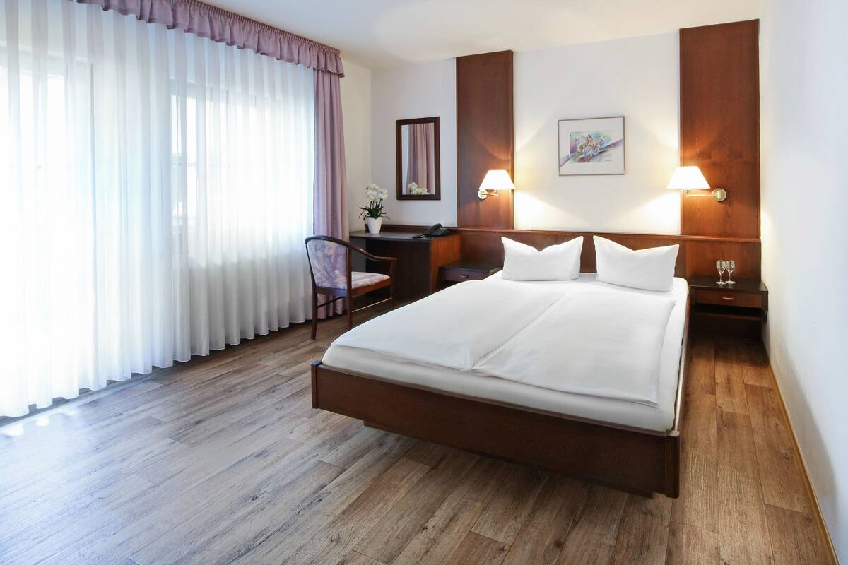 A hotel room with a large bed, a desk, a chair, a lamp, and a window with curtains.