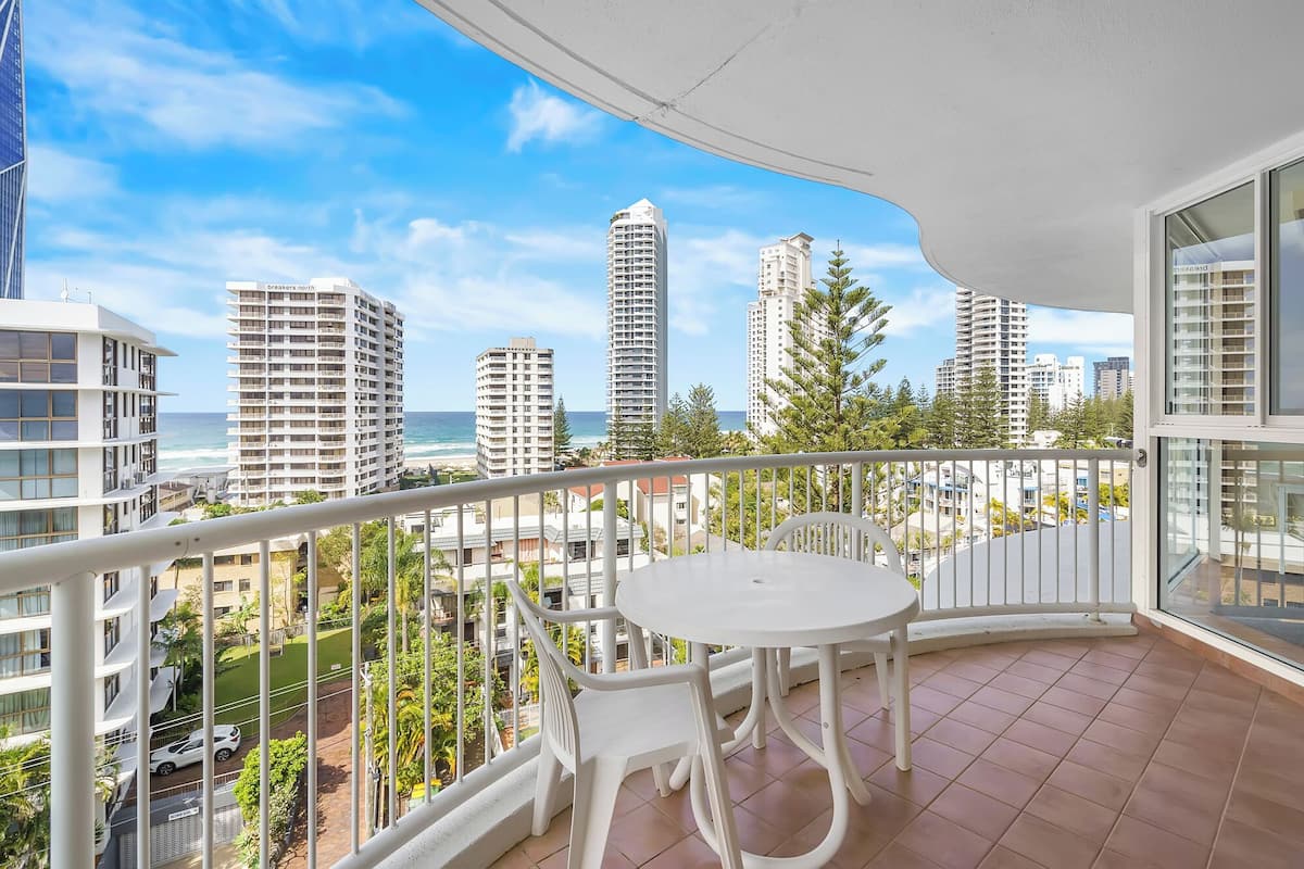 A balcony with a white table and chairs, overlooking a beach and high-rise buildings.