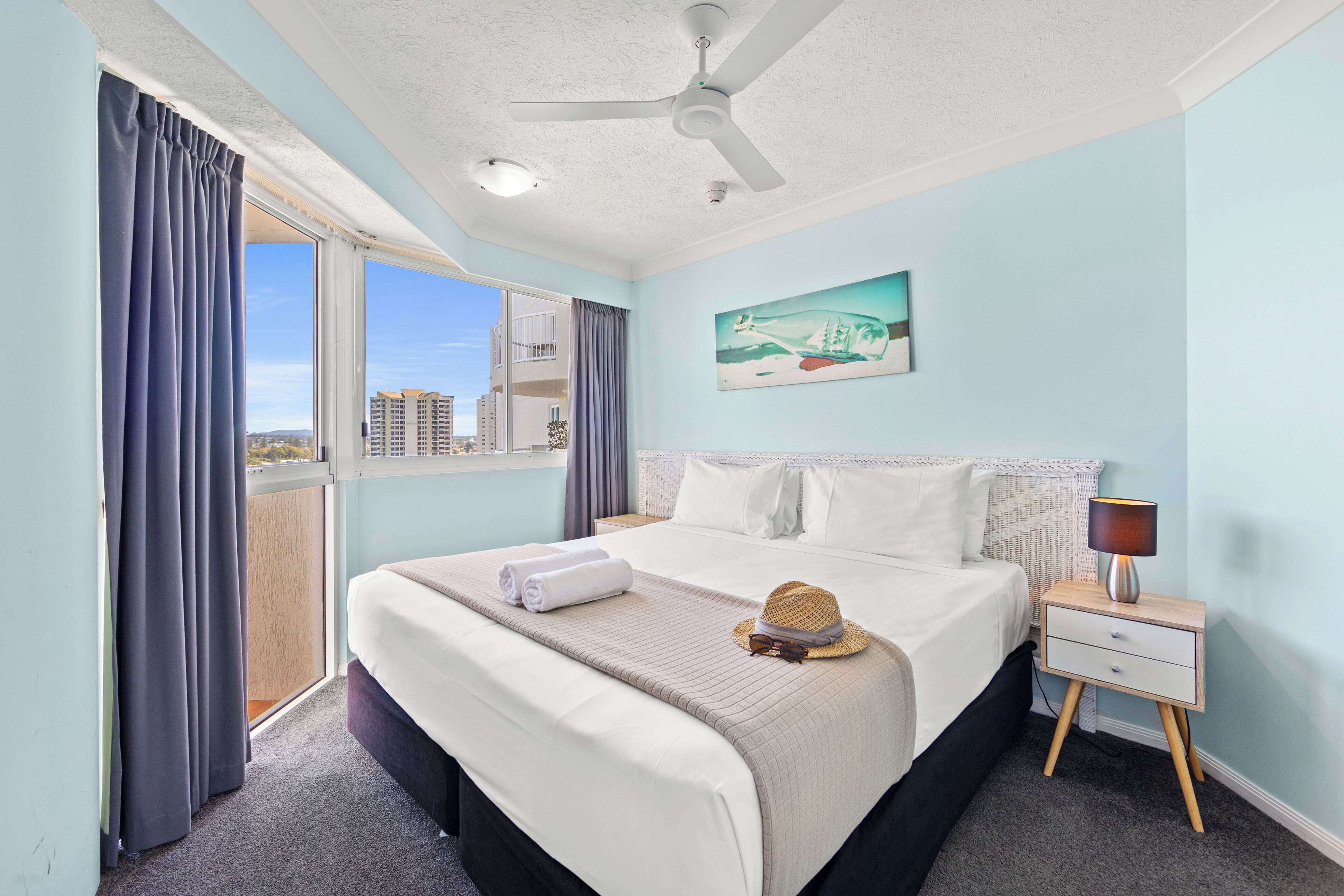 Quarto de hotel com uma cama, mesa de cabeceira, ventoinha no teto, janela com cortinas e um quadro na parede.