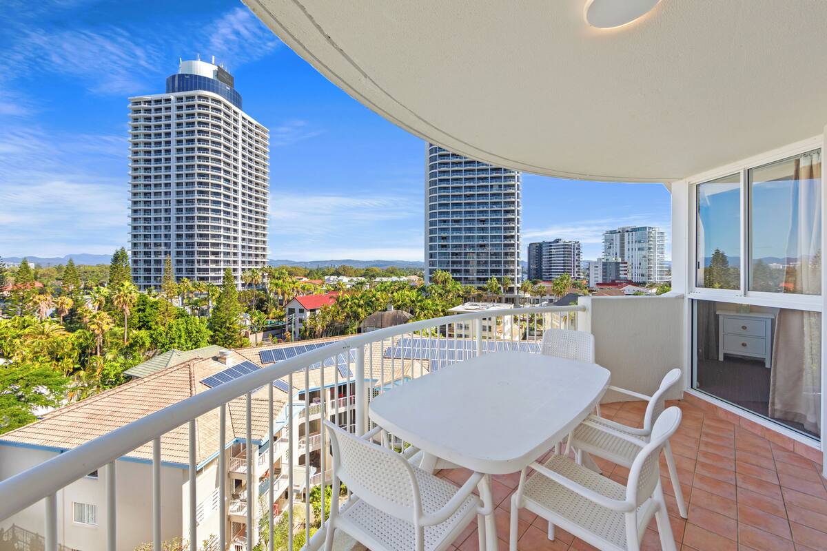 A balcony with a white table and chairs, overlooking a cityscape with tall buildings and greenery.