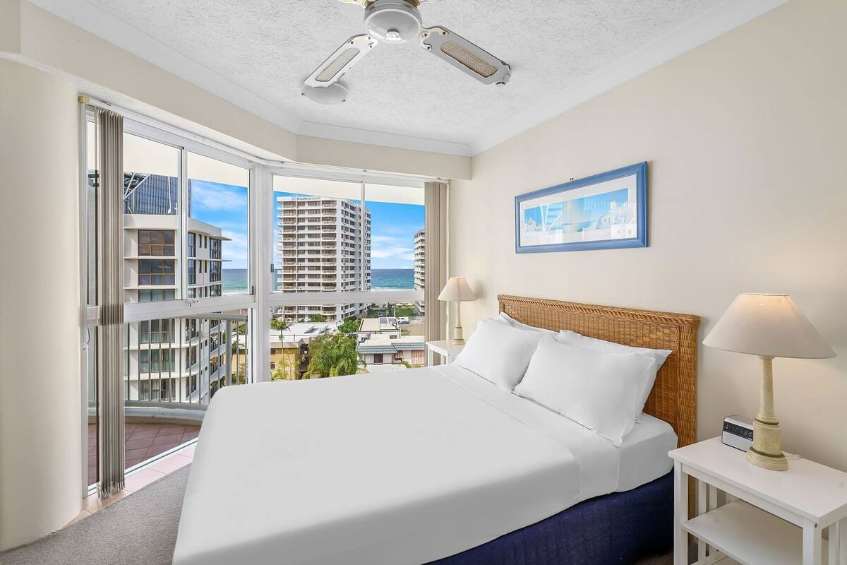 A hotel room with a large bed, a ceiling fan, a view of the ocean, and a framed picture on the wall.