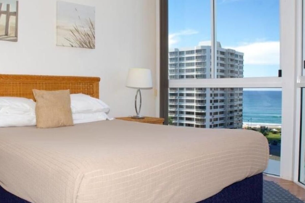 A hotel room with a large bed, a nightstand, a lamp, and a view of a high-rise building and the ocean.