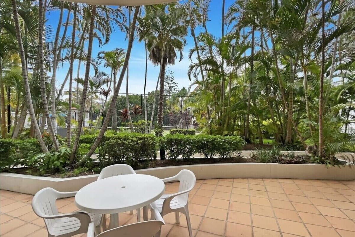 An outdoor patio with a round table and four chairs, surrounded by palm trees and other greenery.