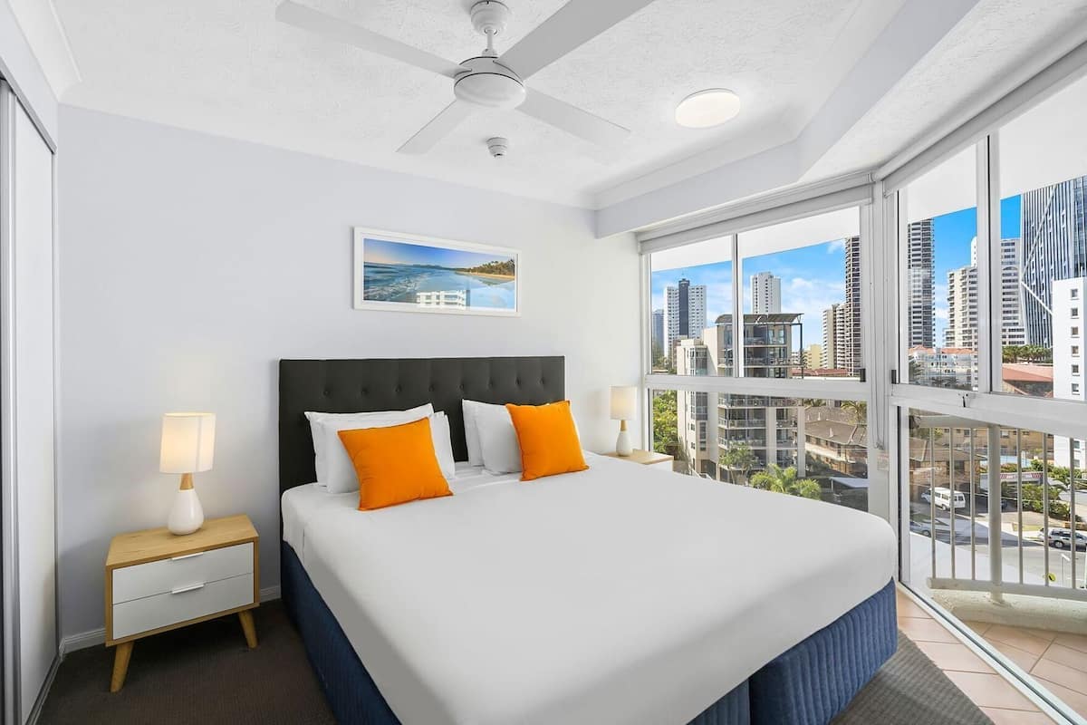 A hotel room with a bed, a nightstand, a lamp, a ceiling fan, and a large window with a city view.