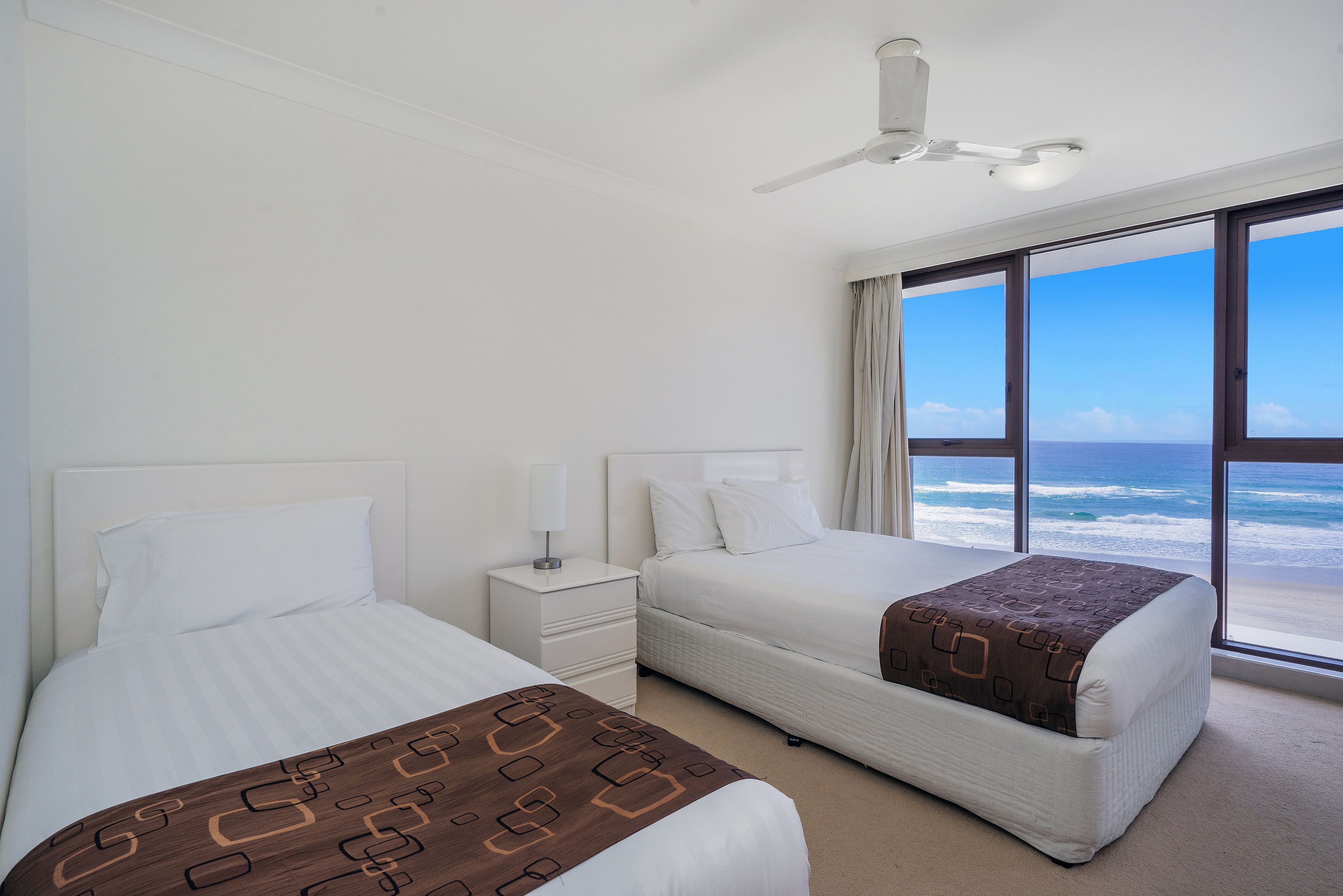 Habitación de hotel con dos camas, un ventilador de techo, una mesita de noche y un ventanal con vista al océano.