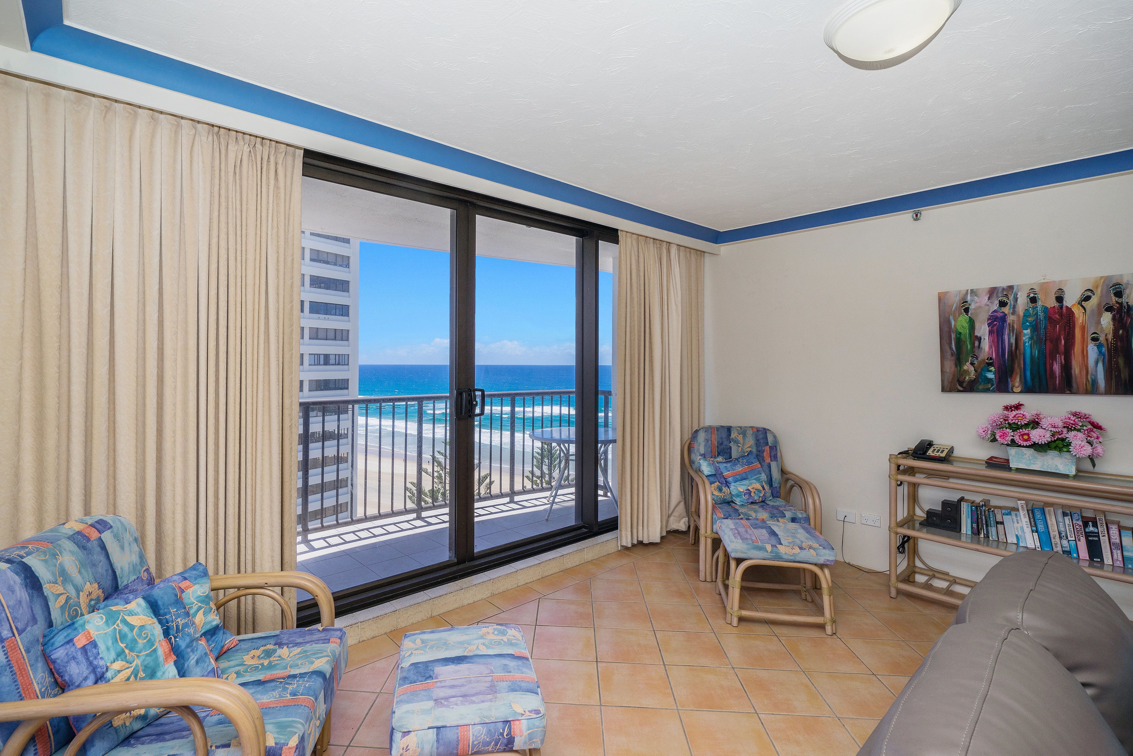 A balcony with a view of the ocean, a sofa, a chair, and a bookshelf with books and flowers.