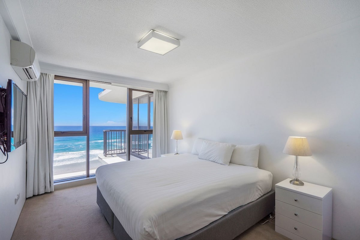 Habitación de hotel con cama, lámparas de noche, mesita de noche, televisión y balcón con vista al océano.
