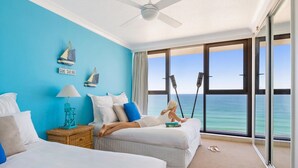 Habitación de hotel con dos camas, un ventanal con vistas al océano, un ventilador de techo y una pequeña mesa con una lámpara.