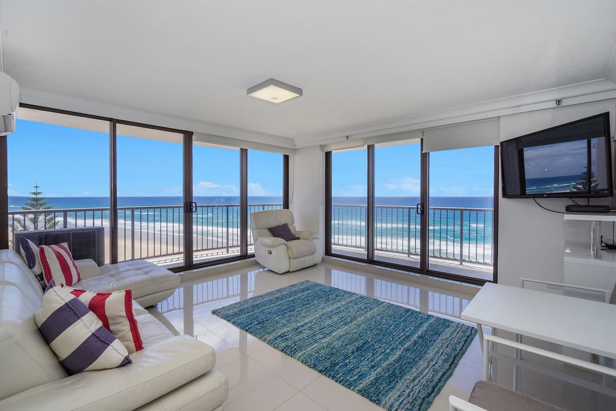 Amplia sala de estar con un gran ventanal con vista a la playa, un sofá blanco con cojines a rayas y un televisor de pantalla plana montado en la pared.