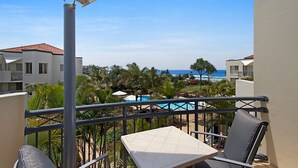 A balcony with a table and chairs, overlooking a pool and palm trees, with buildings and a view of the ocean in the background. - Golden Riviera Beach Resort (Tugun)