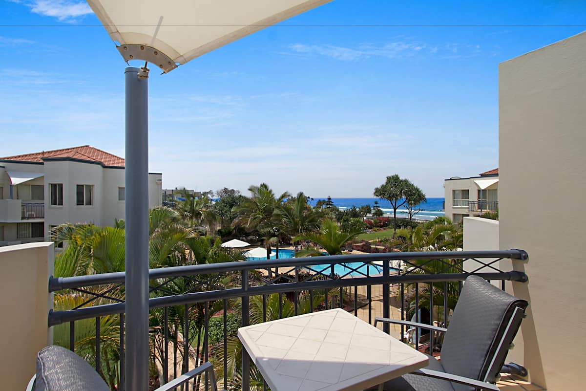 Ein Balkon mit Tisch und Stühlen, Blick auf einen Pool und Palmen, mit Gebäuden und Meerblick im Hintergrund.