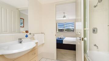 A modern bathroom with a large sink, a shower, and a bed visible through an open door.