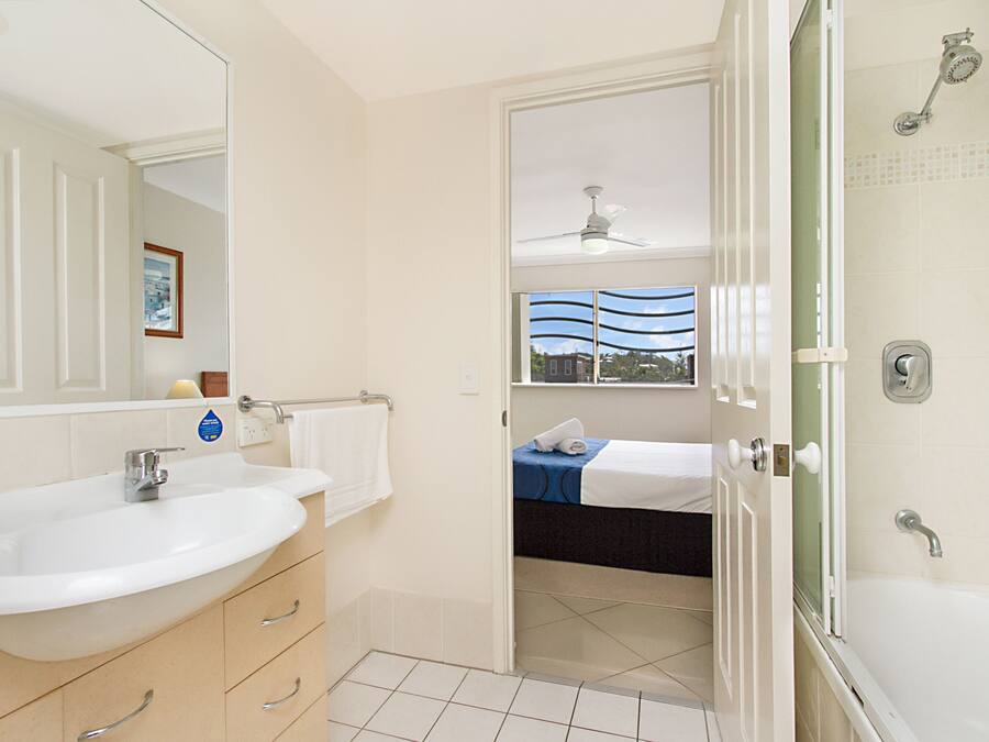 A modern bathroom with a large sink, a shower, and a bed visible through an open door.
