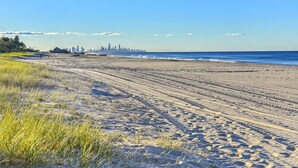 A sandy beach with footprints and sparse grass, leading to a city skyline in the distance. - Golden Riviera Beach Resort (Tugun)