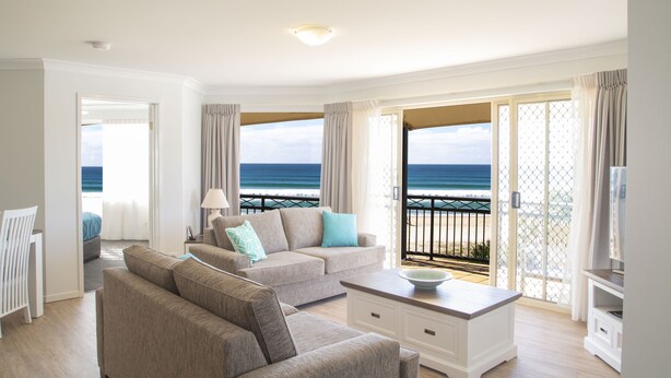 A modern living room with a sofa, a coffee table, and a TV. There is a balcony with a view of the ocean. - Golden Riviera Beach Resort (Tugun)