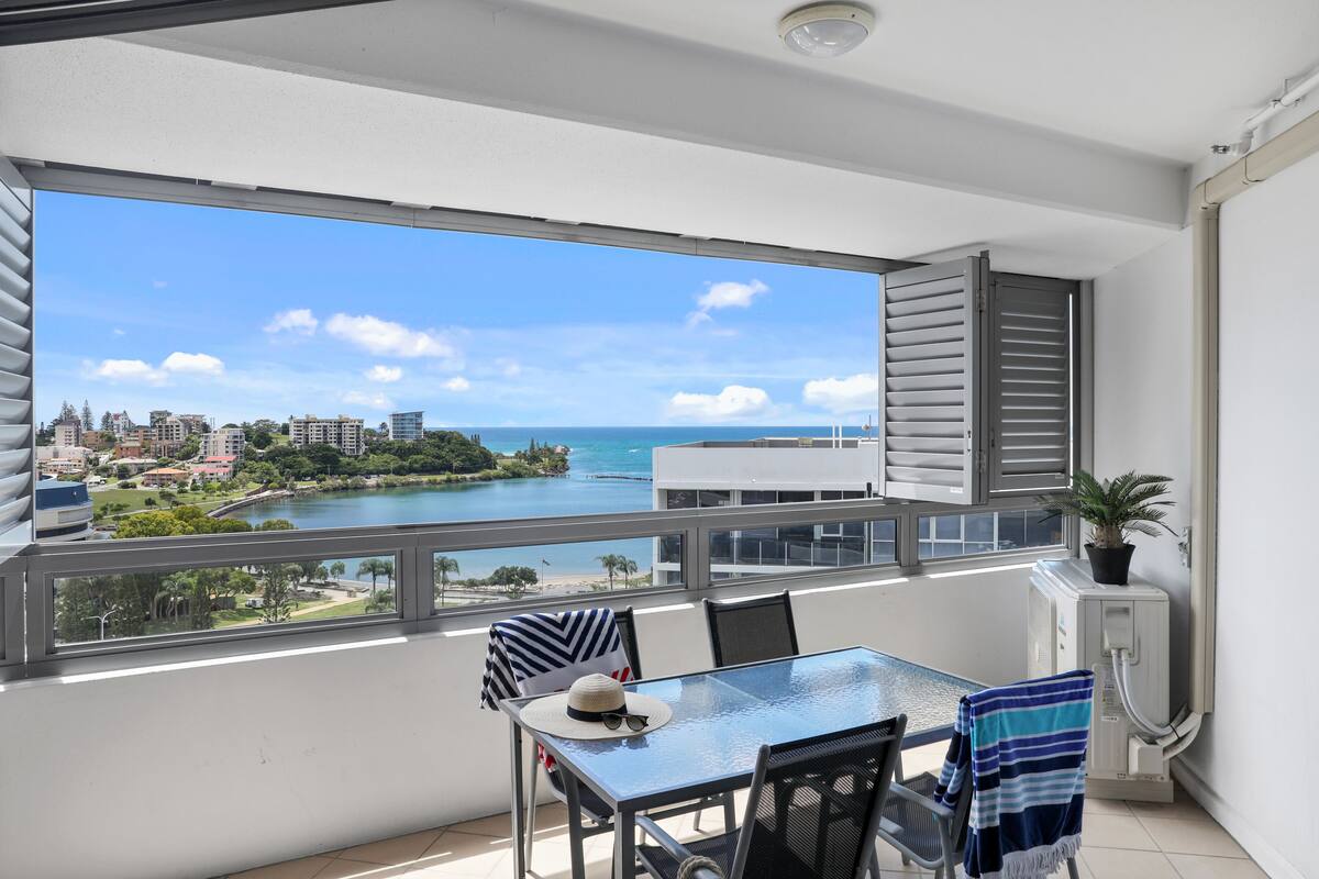 A balcony with a view of a cityscape and a body of water. There is a table with chairs and a potted plant.