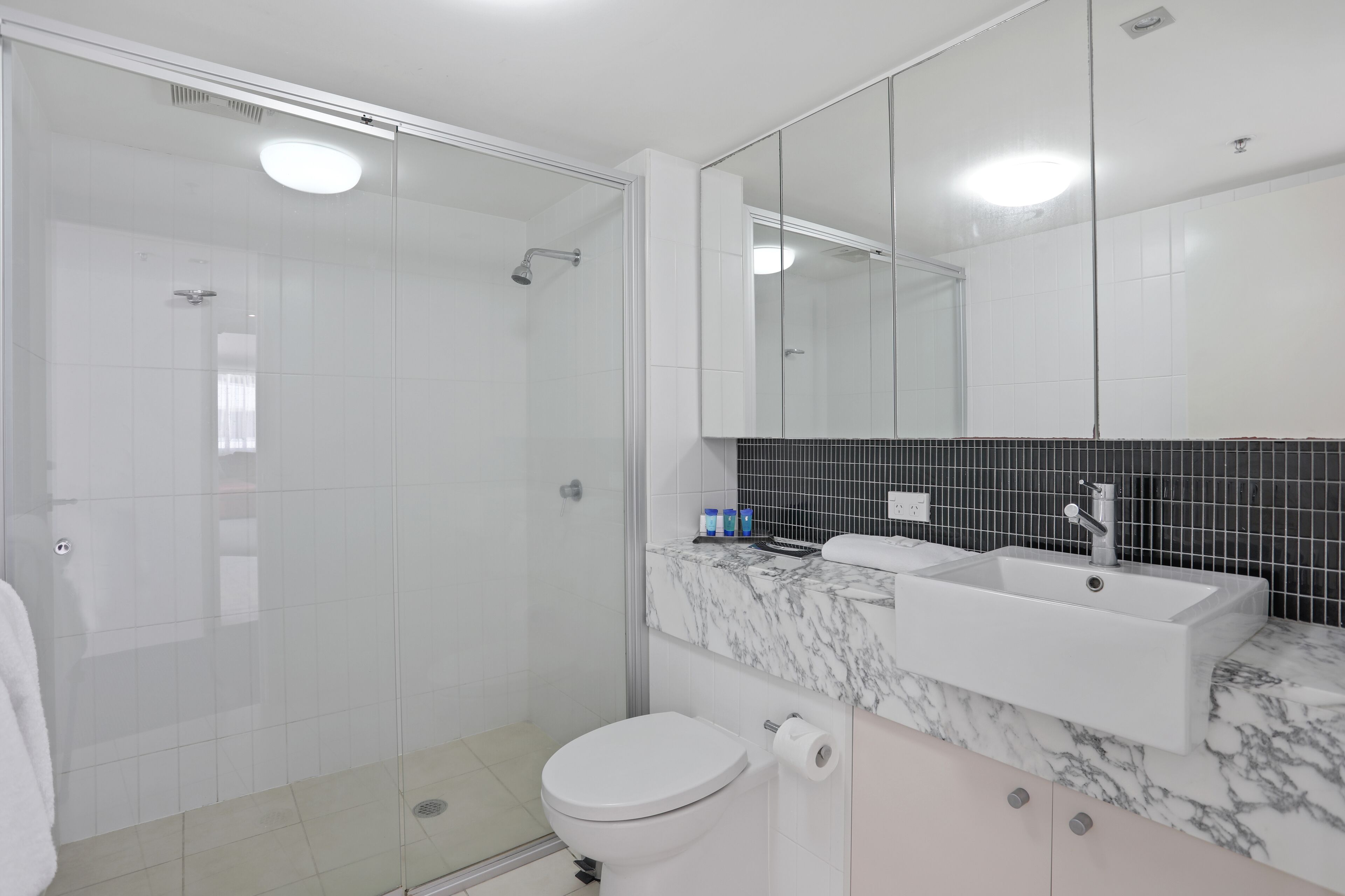 A modern bathroom with a glass-enclosed shower, a white toilet, a sink with a marble countertop, and a large mirror.