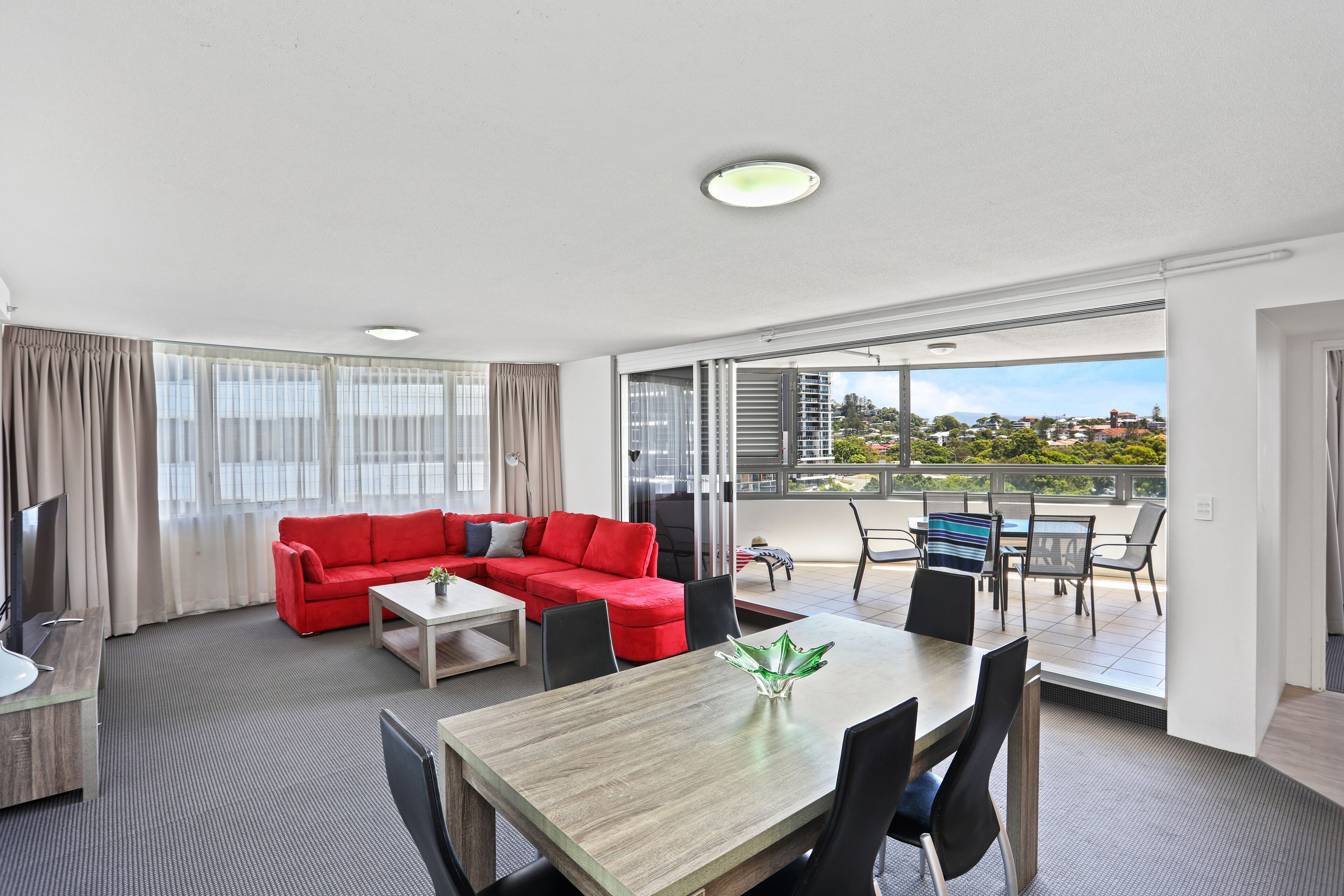 Una habitación de hotel moderna con un sofá rojo, una mesa de comedor y un balcón con vistas a la ciudad.