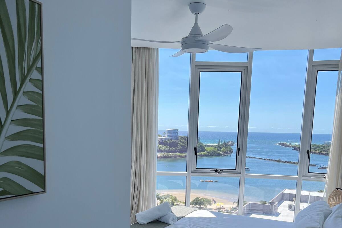 A bedroom with a large bed, a ceiling fan, and a view of the ocean and buildings.