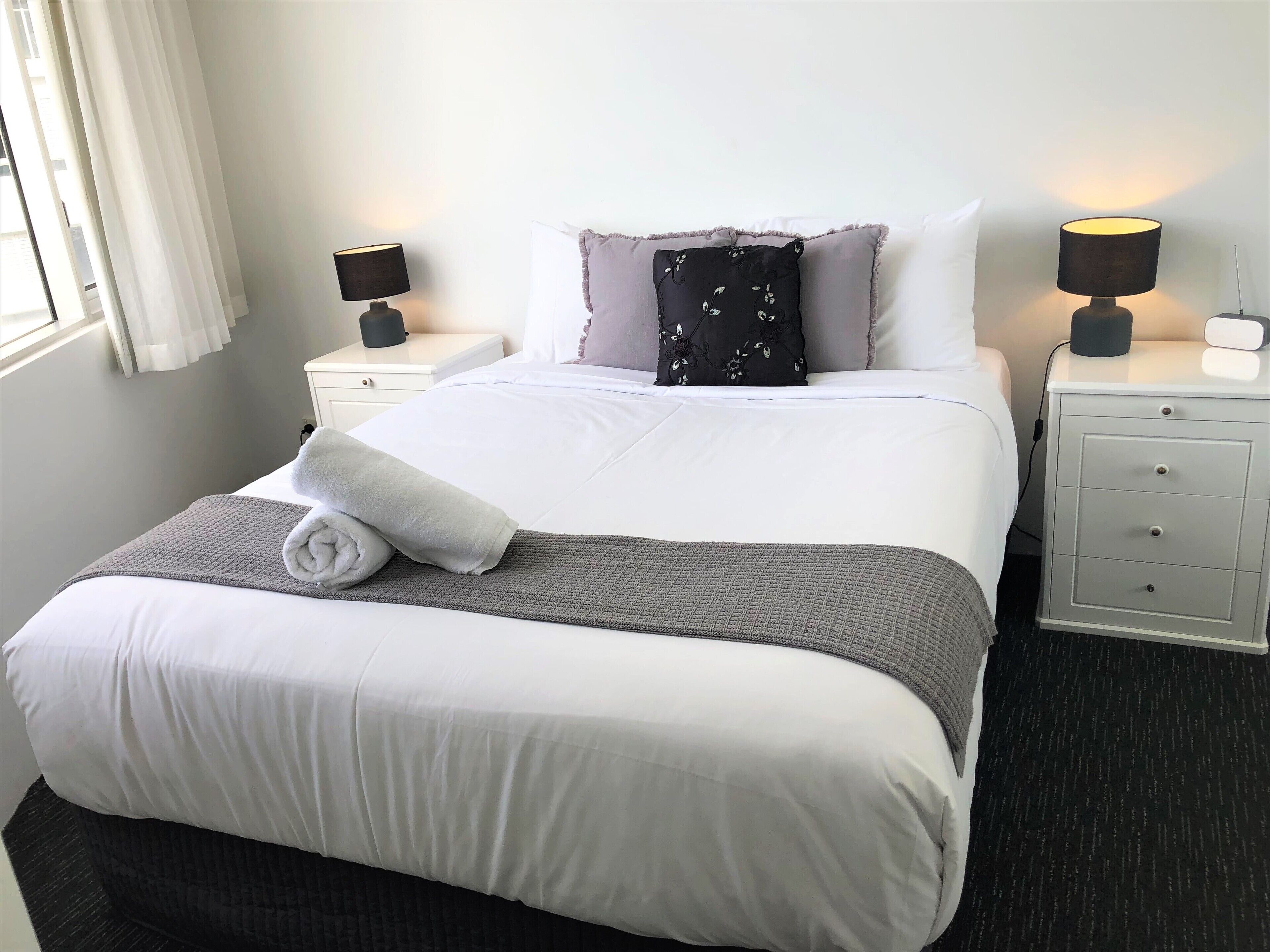 A neatly made bed with a white comforter, a gray throw blanket, and a rolled white towel. There are two bedside tables with lamps, a gray nightstand, and a white wall with a window and curtains.
