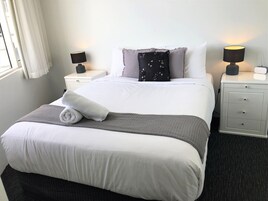 Una cama bien hecha con una colcha blanca, una manta gris y una toalla blanca enrollada. Hay dos mesitas de noche con lámparas, un revistero gris y una pared blanca con una ventana y cortinas.