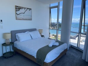 A bedroom with a large bed, two bedside tables with lamps, a view of the ocean through large windows, and a painting on the wall.