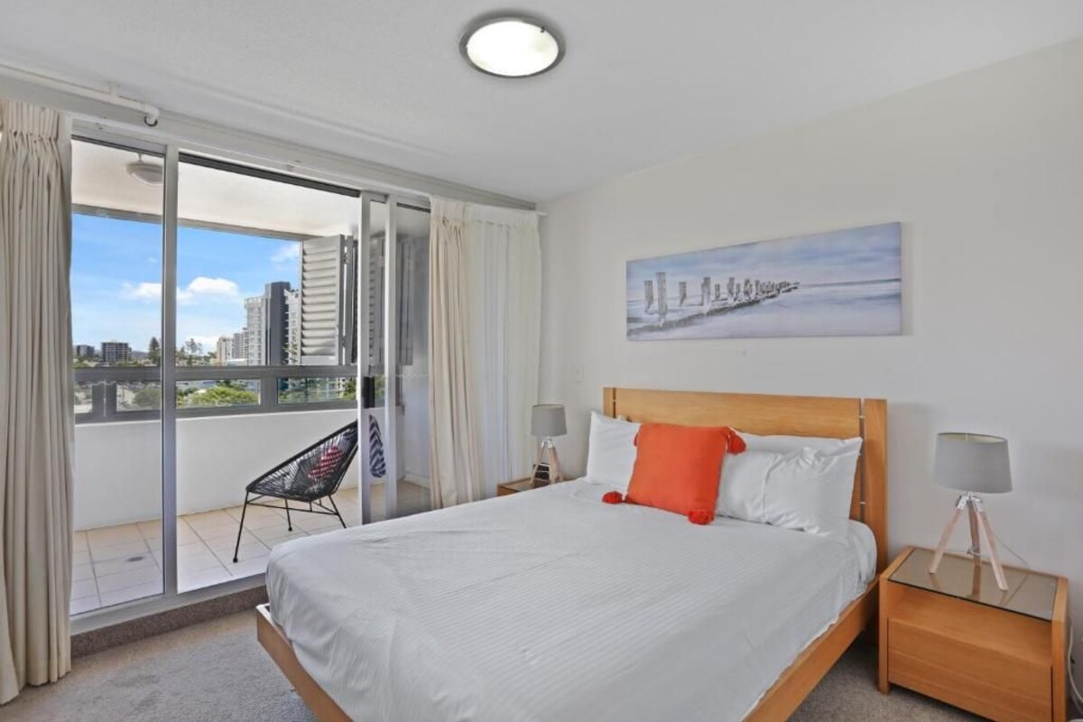A hotel room with a large bed, a balcony with a chair, a painting on the wall, and a city view.