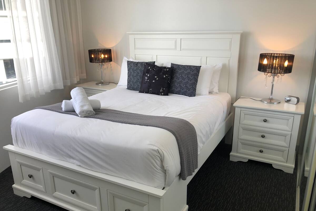 A neatly made bed with white linens, a grey throw, and patterned pillows. There are two bedside tables with lamps, a white headboard, and a dark carpeted floor.