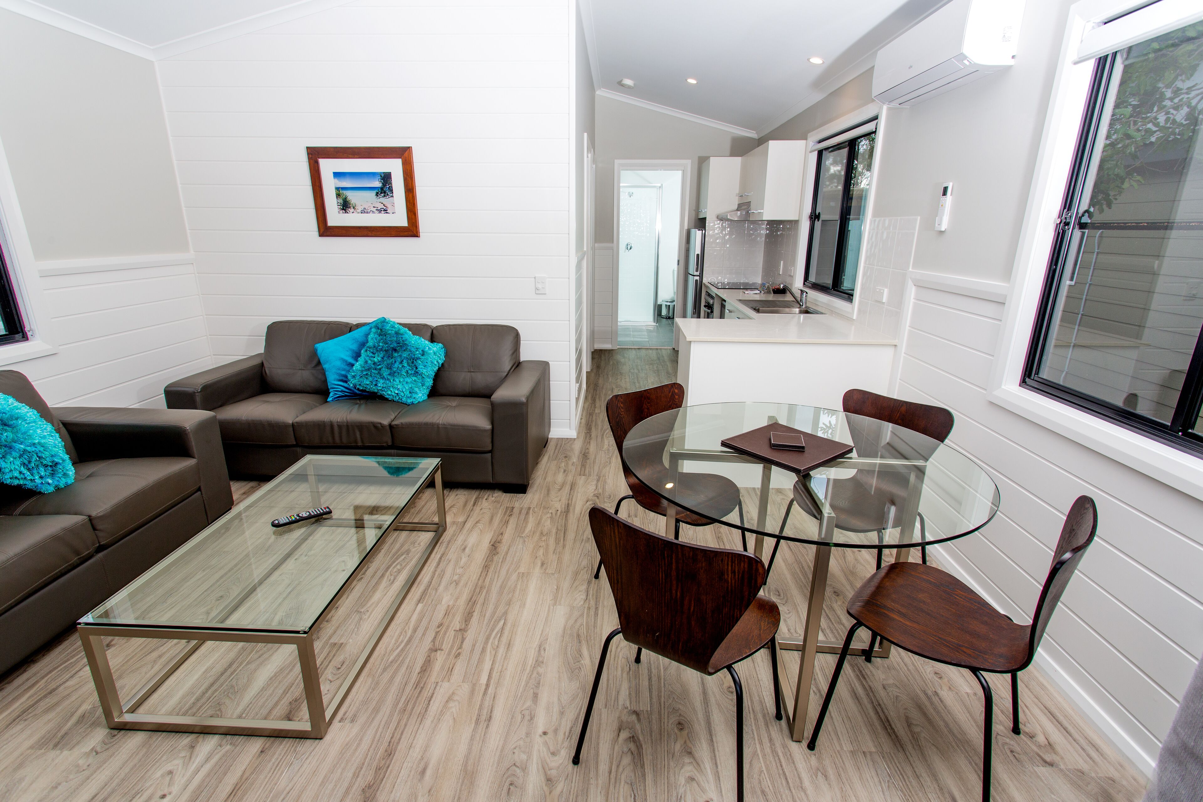 A modern living room with a glass coffee table, brown leather sofa, and teal throw pillows. A dining area with a glass table and wooden chairs is adjacent. The room features white paneled walls and a view into the kitchen through an open doorway.