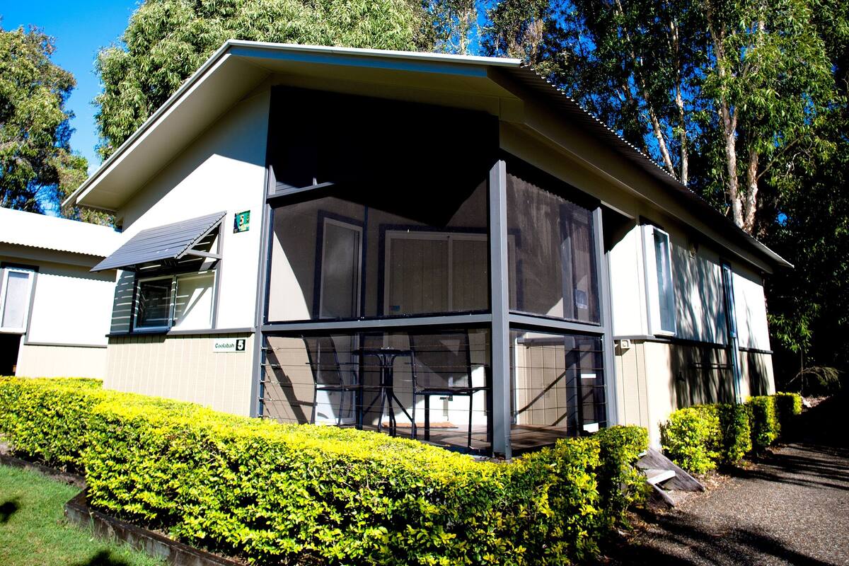 A modern building with a covered outdoor seating area, a visible entrance, and a well-maintained hedge in front.